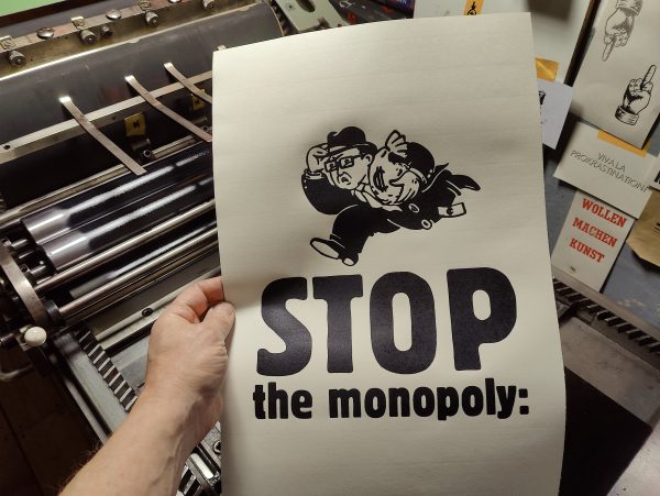 Foto Hand hält einen Abzug vor die Druckmaschine. Der Abzug zeigt im Comic Stil einen Mann mit Homburger Hut, Hornbrille, feistem Gesicht, im Anzug einen Geldsack haltend rennen. Darunter das Wort STOP, darunter die Zeile the monopoly:
