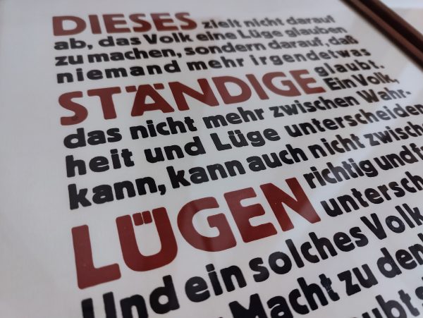 Foto Detail des fertig gerahmten Druckes in Bordaux Rot und Schwarz Blau eines Textes von Hannah Arendt. Der Text lautet: Dieses ständige Lügen zielt nicht darauf ab, das Volk eine Lüge glauben zu machen, sondern darauf, dass niemand mehr irgendetwas glaubt. Ein Volk, das nicht mehr zwischen Wahrheit und Lüge unterscheiden kann, kann auch nicht zwischen richtig und falsch unterscheiden. Und ein solches Volk, das sich seiner Macht, zu denken und zu urteilen, beraubt sieht, ist auch, ohne zu wissen und zu wollen, dem Gesetz der Lüge vollständig unterworfen. Mit einem solchen Volk kann man dann machen, was man will.