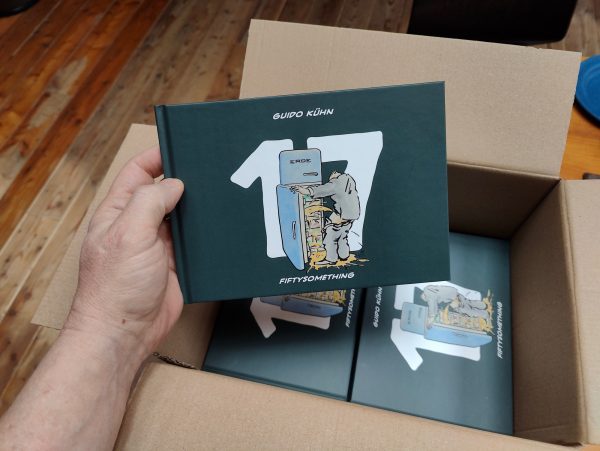 Foto einer Hand, die ein Buch der Ausgabe Fiftysomething 17 aus einem Versandkarton mit anderen Büchern derselben Ausgabe entnimmt.