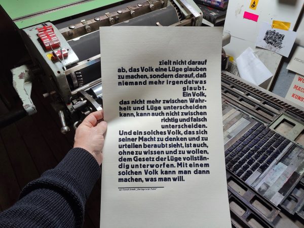 Foto Hand hält die Druckfahne zur Anmutung für die Betrachter*innen vor die Druckmaschine
