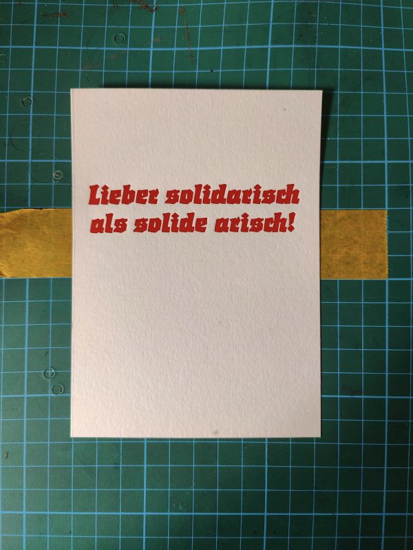 Foto einer Karte in rotem Bleisatz. Der Text der Karte: Lieber solidarisch als solide arisch! Gedruckt in der gebrochenen Werbeschrift Start von Auspurg, erschienen 1935 bei Weber.