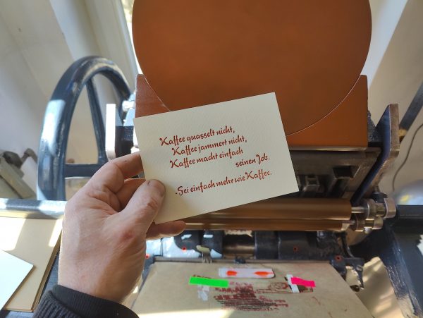 Foto meiner Hand, die eine in braun gedruckte Karte in Bleisatz vor die Druckmaschine hält. Der Text der in einer typischen 1950er Eisdielen und Caféterassen-Werbeschreibschrift ausgeführt ist lautet: Kaffee quasselt nicht, Kaffee jammert nicht, Kaffee macht einfach seinen Job. -Sei einfach mehr wie Kaffee.