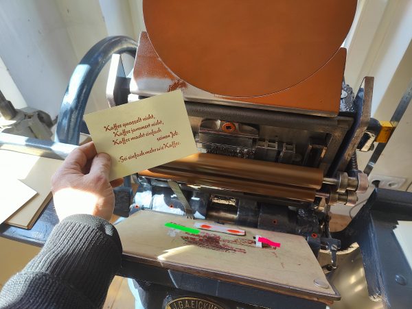 Foto meiner Hand, die eine in braun gedruckte Karte in Bleisatz vor die Druckmaschine hält. Der Text der in einer typischen 1950er Eisdielen und Caféterassen-Werbeschreibschrift ausgeführt ist lautet: Kaffee quasselt nicht, Kaffee jammert nicht, Kaffee macht einfach seinen Job. -Sei einfach mehr wie Kaffee.