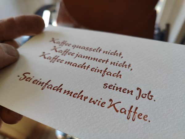 Foto meiner Hand, die eine in braun gedruckte Karte in Bleisatz vor die Druckmaschine hält. Der Text der in einer typischen 1950er Eisdielen und Caféterassen-Werbeschreibschrift ausgeführt ist lautet: Kaffee quasselt nicht, Kaffee jammert nicht, Kaffee macht einfach seinen Job. -Sei einfach mehr wie Kaffee.