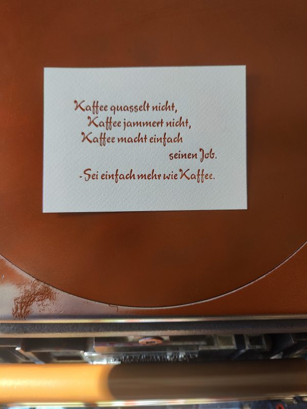 Foto meiner Hand, die eine in braun gedruckte Karte in Bleisatz vor die Druckmaschine hält. Der Text der in einer typischen 1950er Eisdielen und Caféterassen-Werbeschreibschrift ausgeführt ist lautet: Kaffee quasselt nicht, Kaffee jammert nicht, Kaffee macht einfach seinen Job. -Sei einfach mehr wie Kaffee.