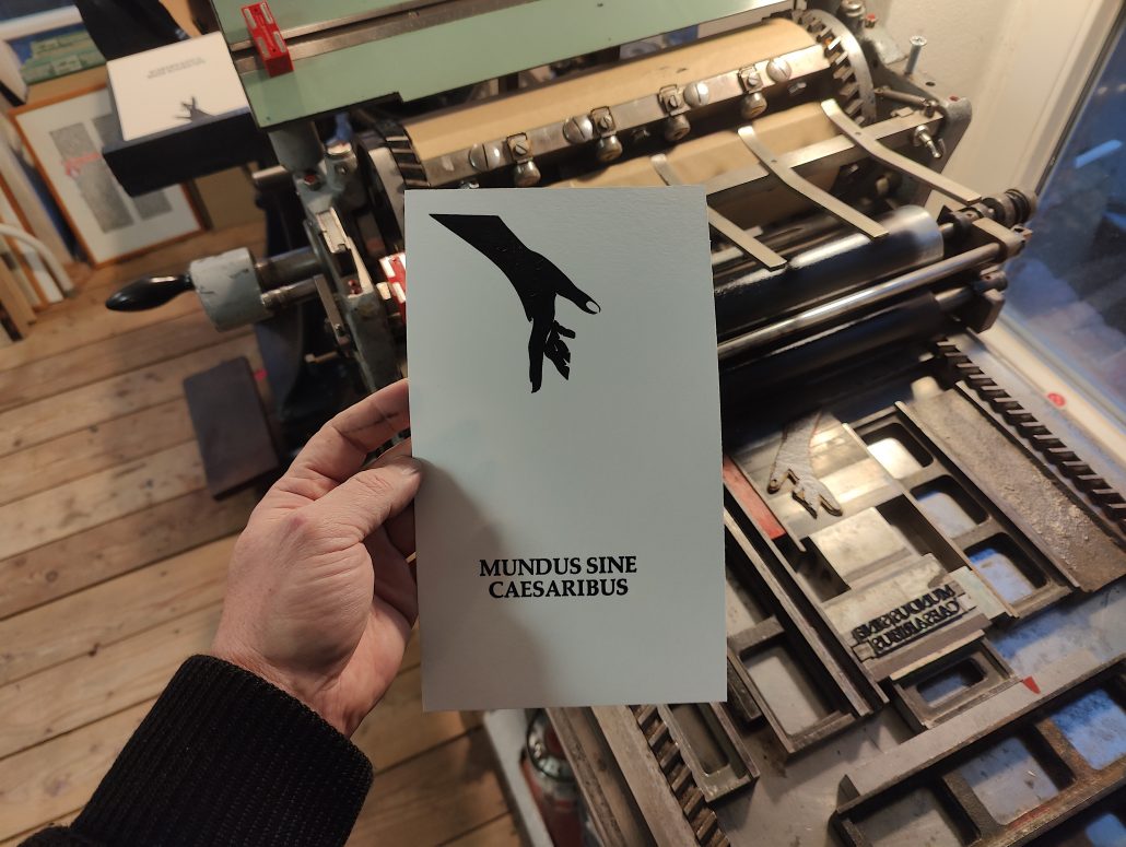 Foto: Hand hält Karte vor Druckmaschine. Zu sehen ist die erste färbe eines zweifarbigen Motivs - eine einen über den Handrücken nach unten geöffnete Hand und der Schriftzug Mundus Sine Caesarius. 