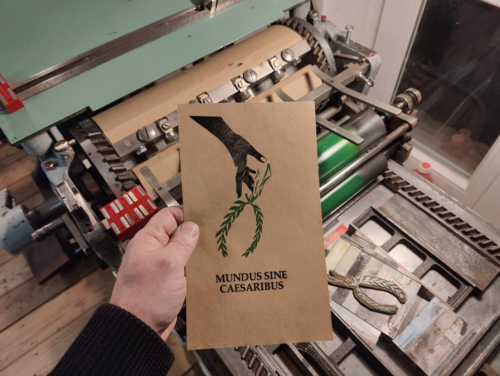 Foto: Hand hält Karte vor Druckmaschine. Zu sehen ist ein zweifarbiger Abzug - eine einen über den Handrücken nach unten geöffnete Hand, die einen Lorbeerkranz fallen lässt über dem Schriftzug Mundus Sine Caesarius. Der Abzug ist auf Makulaturpapier zum einrichten der zweiten Form auf die ersten Farben. 