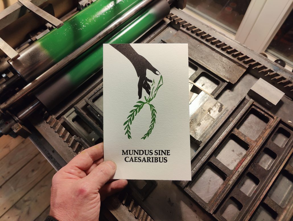 Foto: Hand hält Karte vor Druckmaschine. Zu sehen ist ein zweifarbiger Abzug - eine einen über den Handrücken nach unten geöffnete Hand, die einen Lorbeerkranz fallen lässt über dem Schriftzug Mundus Sine Caesarius. Der Abzug ist auf Baumwollpapier. 