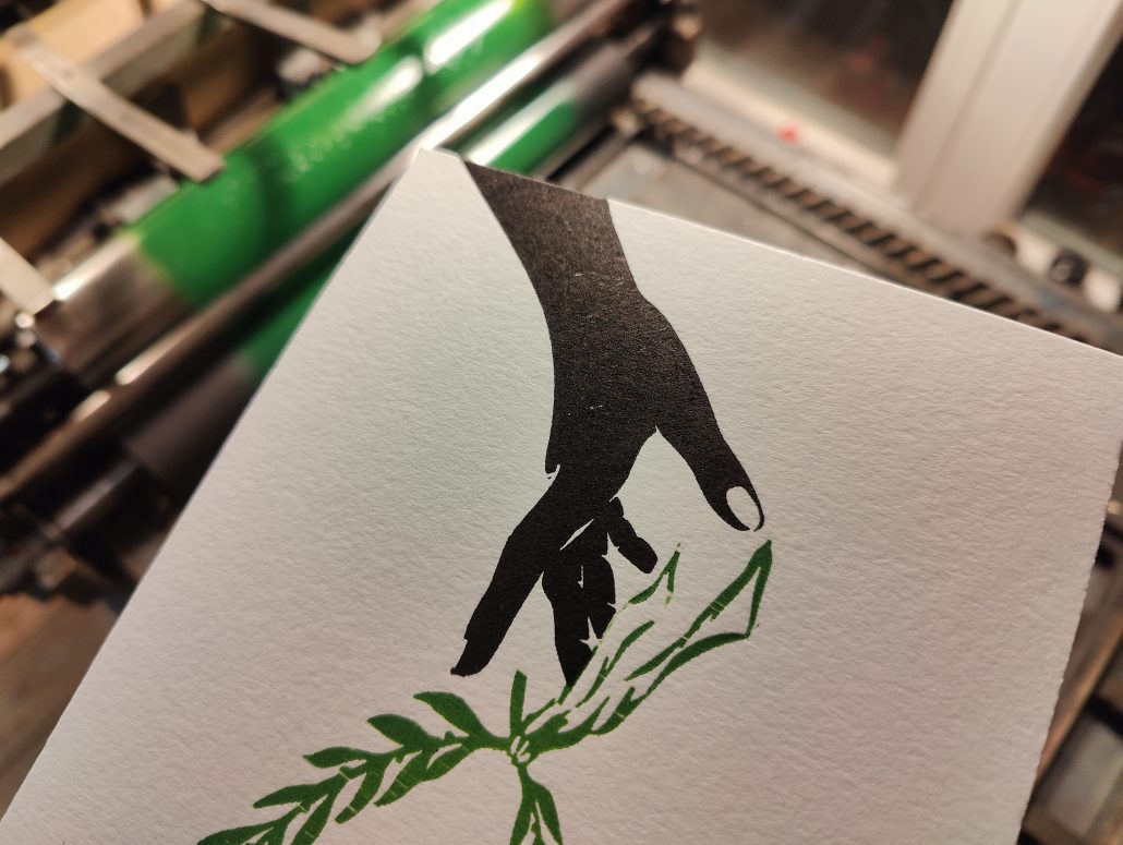 Foto: Hand hält Karte vor Druckmaschine. Zu sehen ist ein zweifarbiger Abzug - eine einen über den Handrücken nach unten geöffnete Hand, die einen Lorbeerkranz fallen lässt.