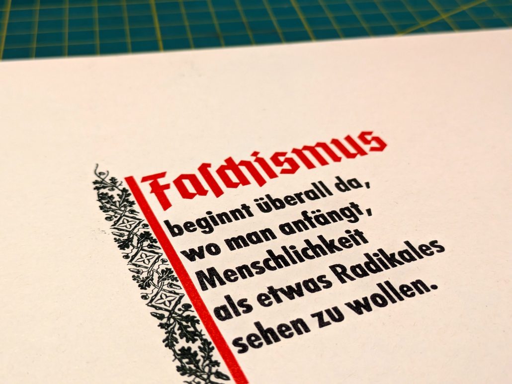Foto Detail des Kopfstückens eines dreifarbigen Druckes. Fahnenartig leicht schräg gestellt sieht man eine rote Linie an deren oberen Ende in roter Fraktur rechts anschlagend das Wort Faschismus steht. Darunter formal als Fahnentuch wird der Text in schwarz gedruckter Futura auf fünf Zeilen verteilt fortgesetzt: beginnt überall da, wo man anfängt, Menschlichkeit als etwas Radikales sehen zu wollen. Links neben der roten Flaggenstockline ist in dunklem Grün eine breite Schmucklinie aus nach oben und unten sich verjüngendem Eichenlaub eingedruckt. 