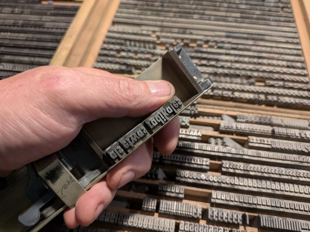 Foto meiner Hand mit Setzerkell über Setzkasten, auf der gerade ein Satz aus dem Setzkasten auf einer Setzlinie arrangiert wird. 