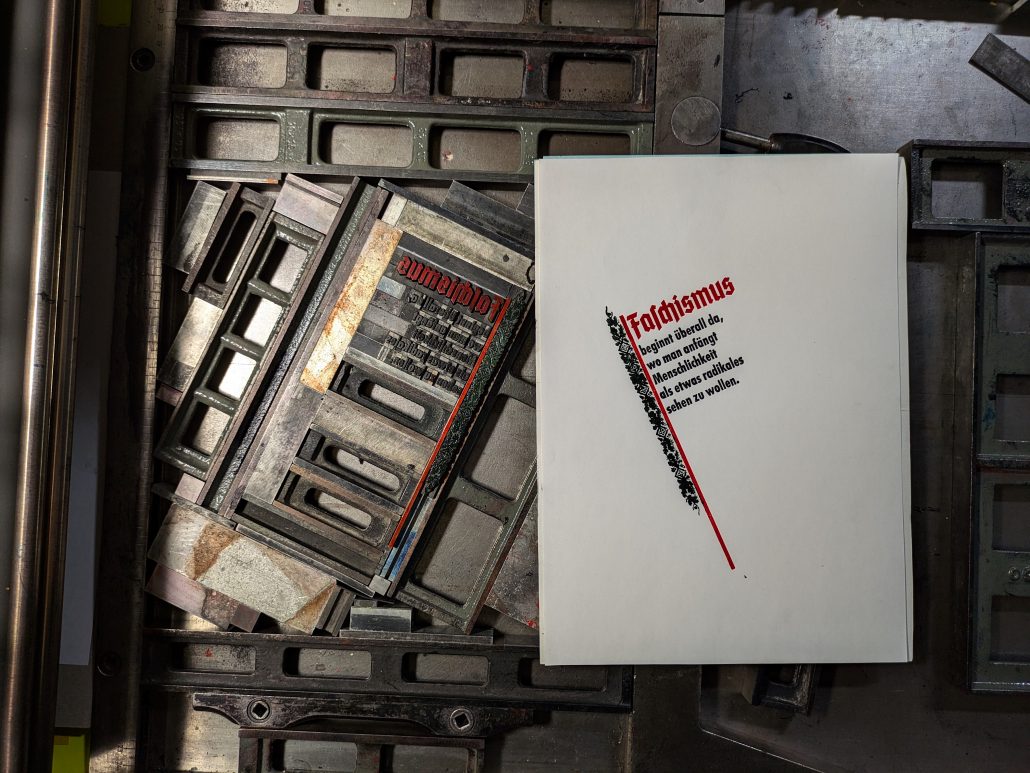 Foto eine dreifarbigen Probedrucks neben der Bleisatzform auf der Druckmaschine. Der fahnenartige Text liest sich: Faschismus beginnt überall da, wo man beginnt Menschlichkeit als etwas Radikales sehen zu wollen. 