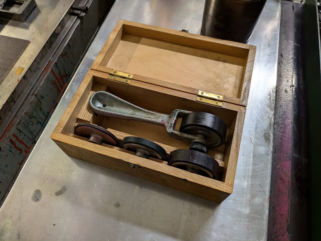 Foto eines Holzköfferchens mit einem Bacher-Linienwalzenset. Das Besteht aus einem Griffstück und verschiedenen, unterschiedlich breiten aber sehr schmalen austauschbaren Farbwalzen. 