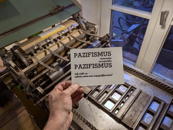Foto Hand hält einen Abzug mit der ersten Farbe, einem tiefblauschwarz vor die Andruckpresse