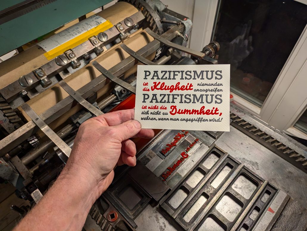 Foto des fertigen, zweifarbigen Drucks in tiefblauschwarz und Pantone 1302 warm-rot auf den Gitterstegen, mit denen die Bleisatzform montiert ist. Der auf sieben Zeil in der Breite des ersten Wortes kompresse (=ohne Durchschuss) gesetzte Text lautet: PAZIFISMUS ist die Klugheit niemanden anzugreifen PAZIFISMUS ist nicht die Dummheit sich nicht zu wehren, wenn man angegriffen wird!