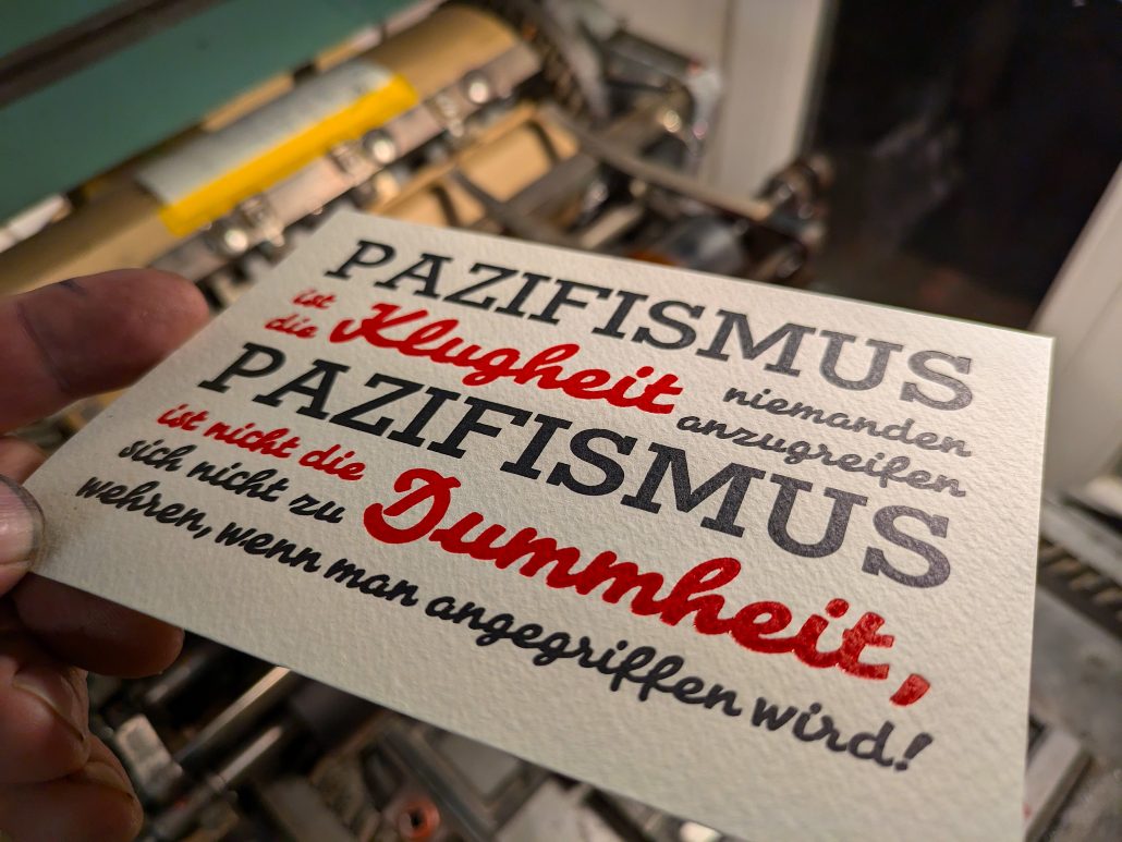 Foto des fertigen, zweifarbigen Drucks in tiefblauschwarz und Pantone 1302 warm-rot auf den Gitterstegen, mit denen die Bleisatzform montiert ist. Der auf sieben Zeil in der Breite des ersten Wortes kompresse (=ohne Durchschuss) gesetzte Text lautet: PAZIFISMUS ist die Klugheit niemanden anzugreifen PAZIFISMUS ist nicht die Dummheit sich nicht zu wehren, wenn man angegriffen wird!