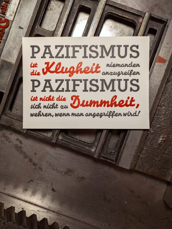 Foto des fertigen, zweifarbigen Drucks in tiefblauschwarz und Pantone 1302 warm-rot auf den Gitterstegen, mit denen die Bleisatzform montiert ist. Der auf sieben Zeil in der Breite des ersten Wortes kompresse (=ohne Durchschuss) gesetzte Text lautet: PAZIFISMUS ist die Klugheit niemanden anzugreifen PAZIFISMUS ist nicht die Dummheit sich nicht zu wehren, wenn man angegriffen wird!