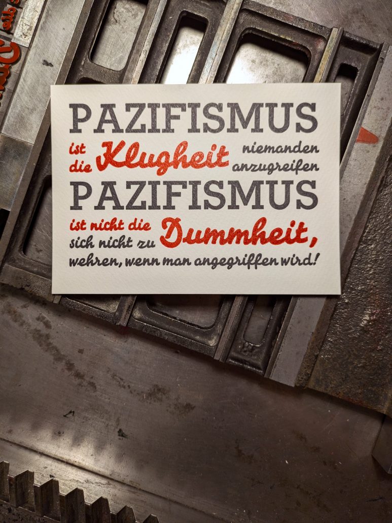 Foto des fertigen, zweifarbigen Drucks in tiefblauschwarz und Pantone 1302 warm-rot auf den Gitterstegen, mit denen die Bleisatzform montiert ist. Der auf sieben Zeil in der Breite des ersten Wortes kompresse (=ohne Durchschuss) gesetzte Text lautet: PAZIFISMUS ist die Klugheit niemanden anzugreifen PAZIFISMUS ist nicht die Dummheit sich nicht zu wehren, wenn man angegriffen wird!