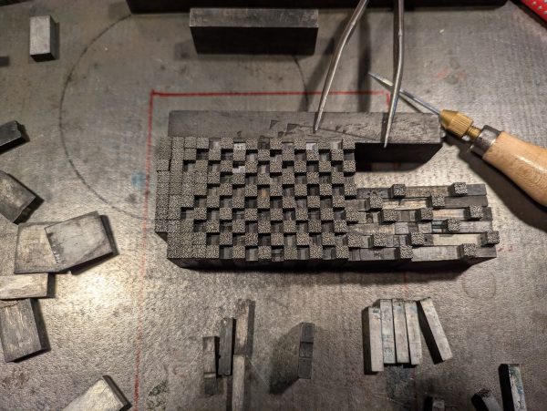 Bleisatzarbeit auf der Schließplatte im entstehen. Man sieht jede Menge Bleisatz-Stempel, welche in der im Enstehen begriffenen Form bereits angeordnet stehen, Blindmaterial und Werkzeug.
