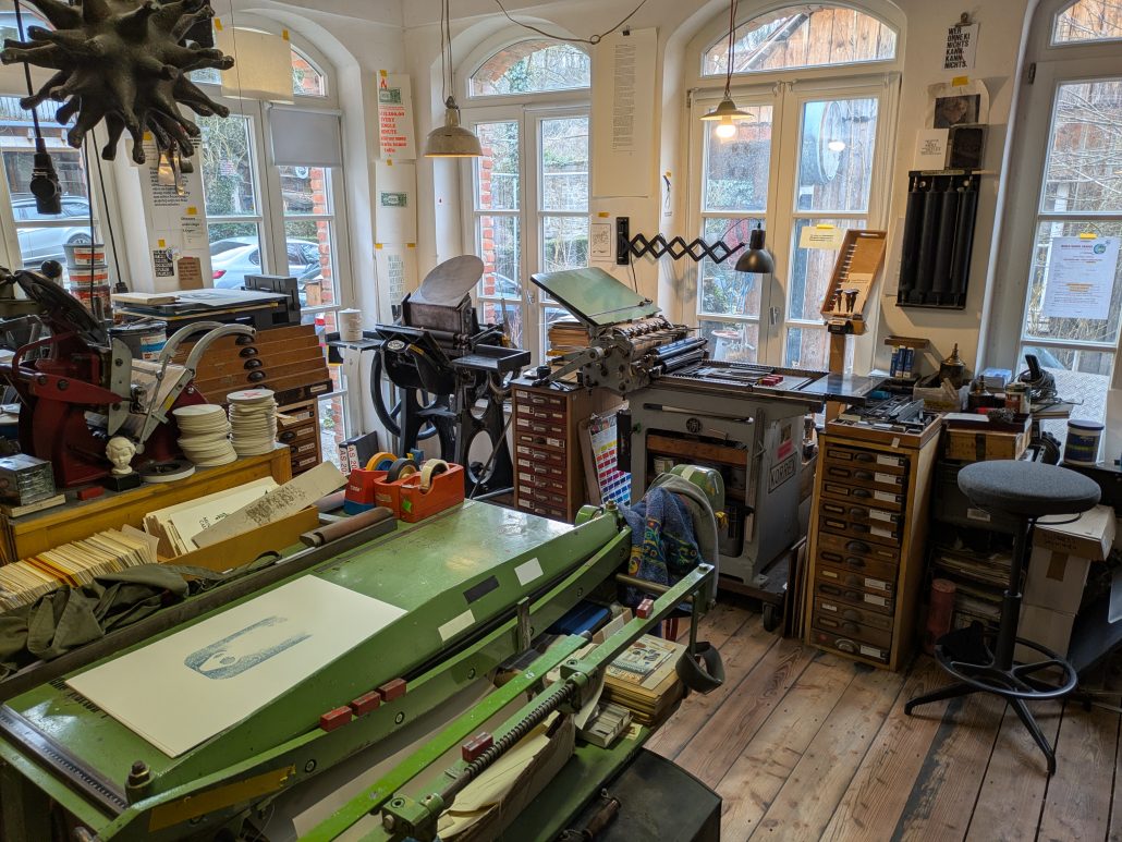 Ein heller, handwerklich geprägter Druckereiraum voller traditioneller Drucktechnik. Mehrere historische Druckpressen stehen dicht beieinander, darunter eine kleine Zylinderpresse und eine zwei Tiegelpressen. Links und rechts reihen sich hohe Setzkästen und Schubladenschränke, gefüllt mit Bleisatz, Holzlettern und Werkzeugen. Auf den Arbeitsflächen liegen Papierstapel, Druckbögen und verschiedene Hilfsmittel. Im Vordergrund steht eine grüne Schneidemaschine, auf deren Fläche frisch gedruckte Blätter liegen. Die Wände sind mit typografischen Postern und Informationsblättern dekoriert. Durch große Rundbogenfenster fällt warmes Tageslicht in den Raum und beleuchtet die etwas chaotisch anmutende, aber sorgfältig organisierte Werkstatt